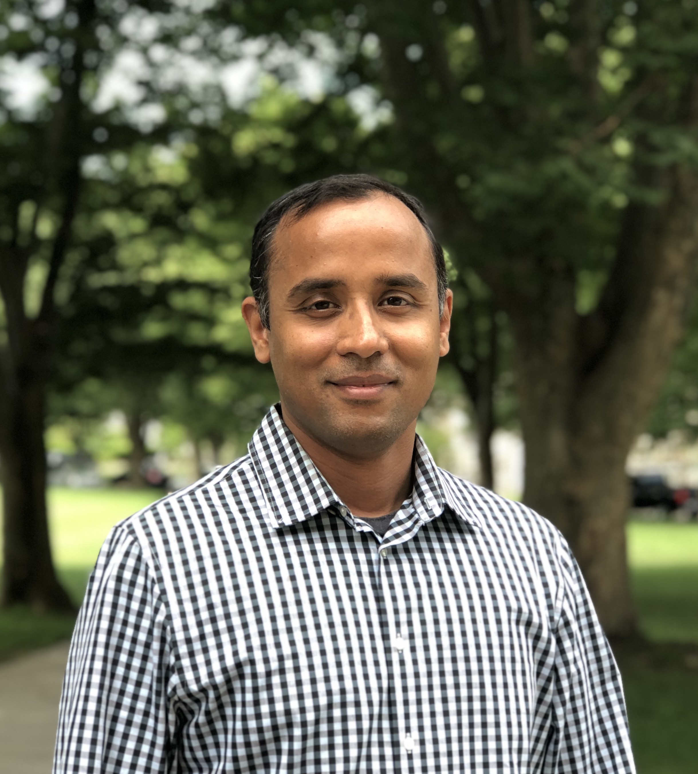 Picture of Krishna Venkatasubramanian in a check button-down shirt. URI quad in the background.